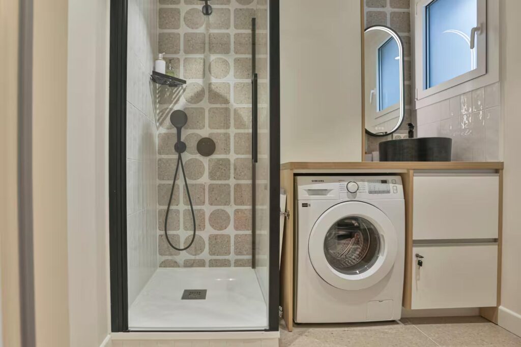 Rénovation de salle d'eau moderne avec douche à l'italienne, carrelage effet pierre et intégration d'un lave-linge sous plan de vasque.