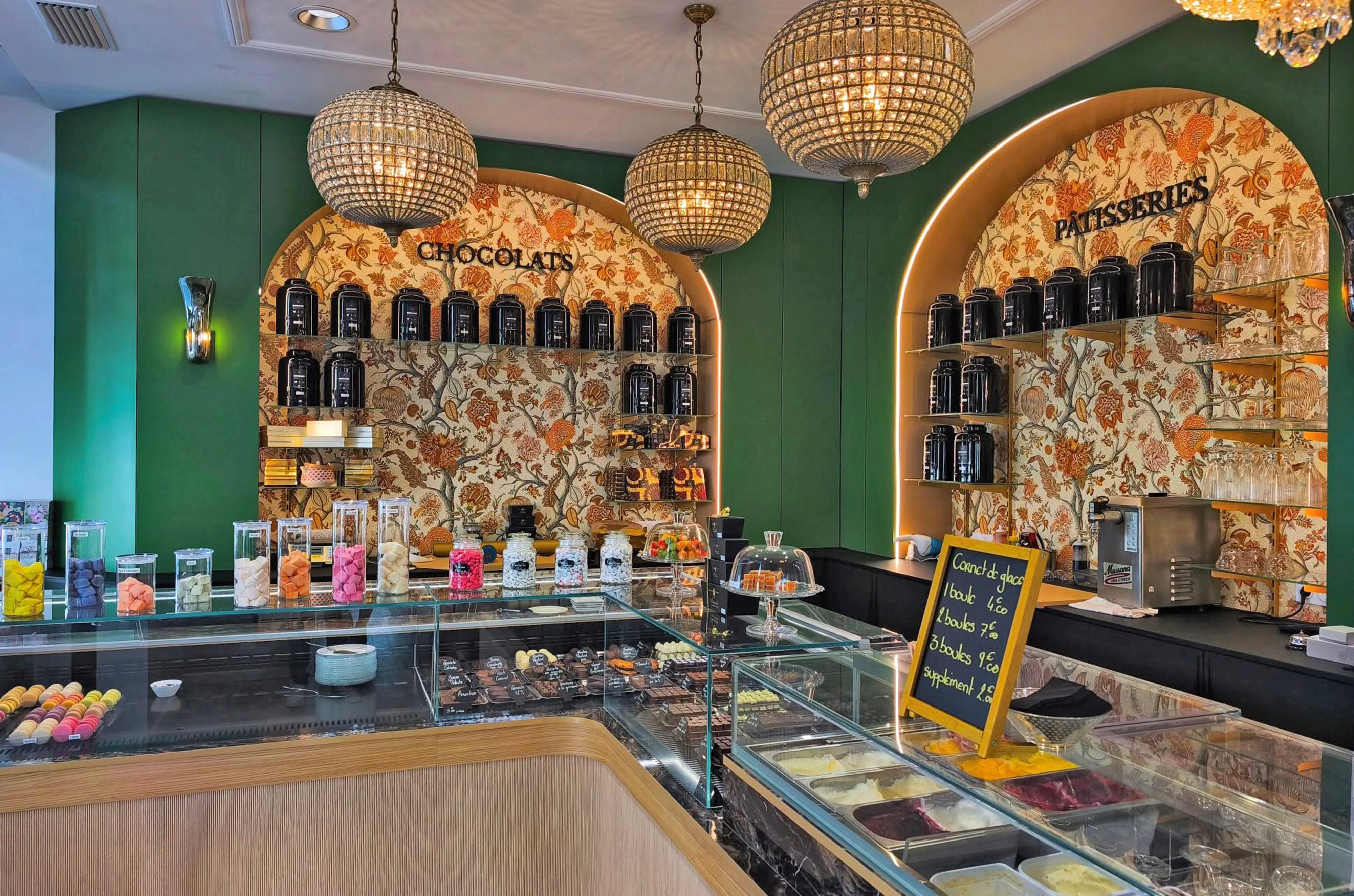 Rayonnages de vente avec papier peint fleuri Pierre Frey, bocaux de confiseries colorées et éclairage par suspensions boules en cristal ciselé.