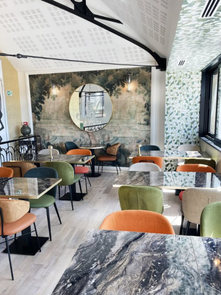 Espace dégustation raffiné avec fauteuils en velours multicolores, tables en marbre noir et papier peint panoramique forestier créant une atmosphère de salon de thé d'autrefois.