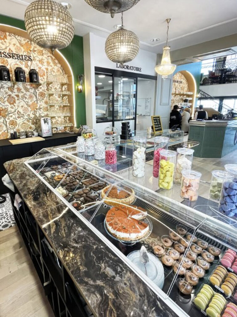 Vitrine réfrigérée moderne présentant une sélection de chocolats artisanaux, avec en arrière-plan le laboratoire de pâtisserie visible à travers une verrière noire.