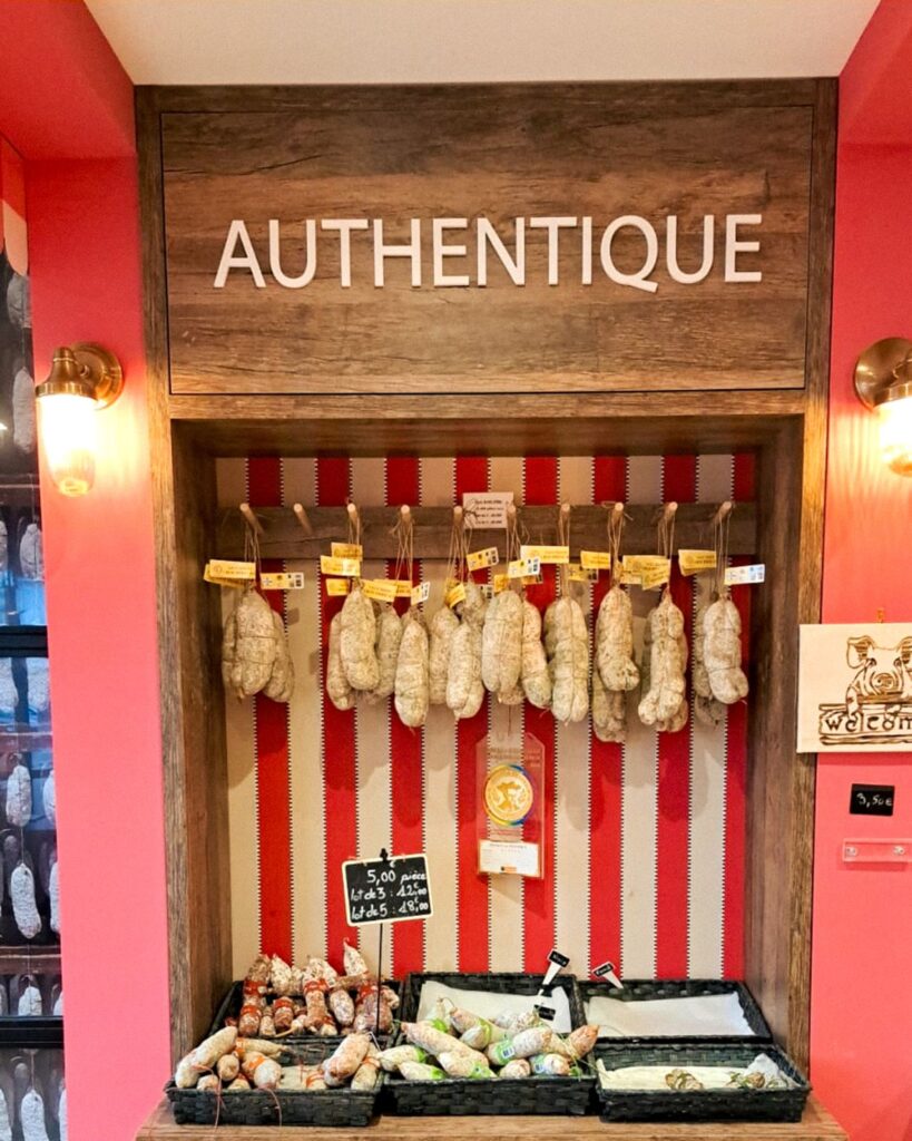 Zoom sur une niche de présentation en bois intitulée "AUTHENTIQUE", mettant en valeur des saucissons artisanaux suspendus sur un fond tapissé de rayures rouges et blanches.