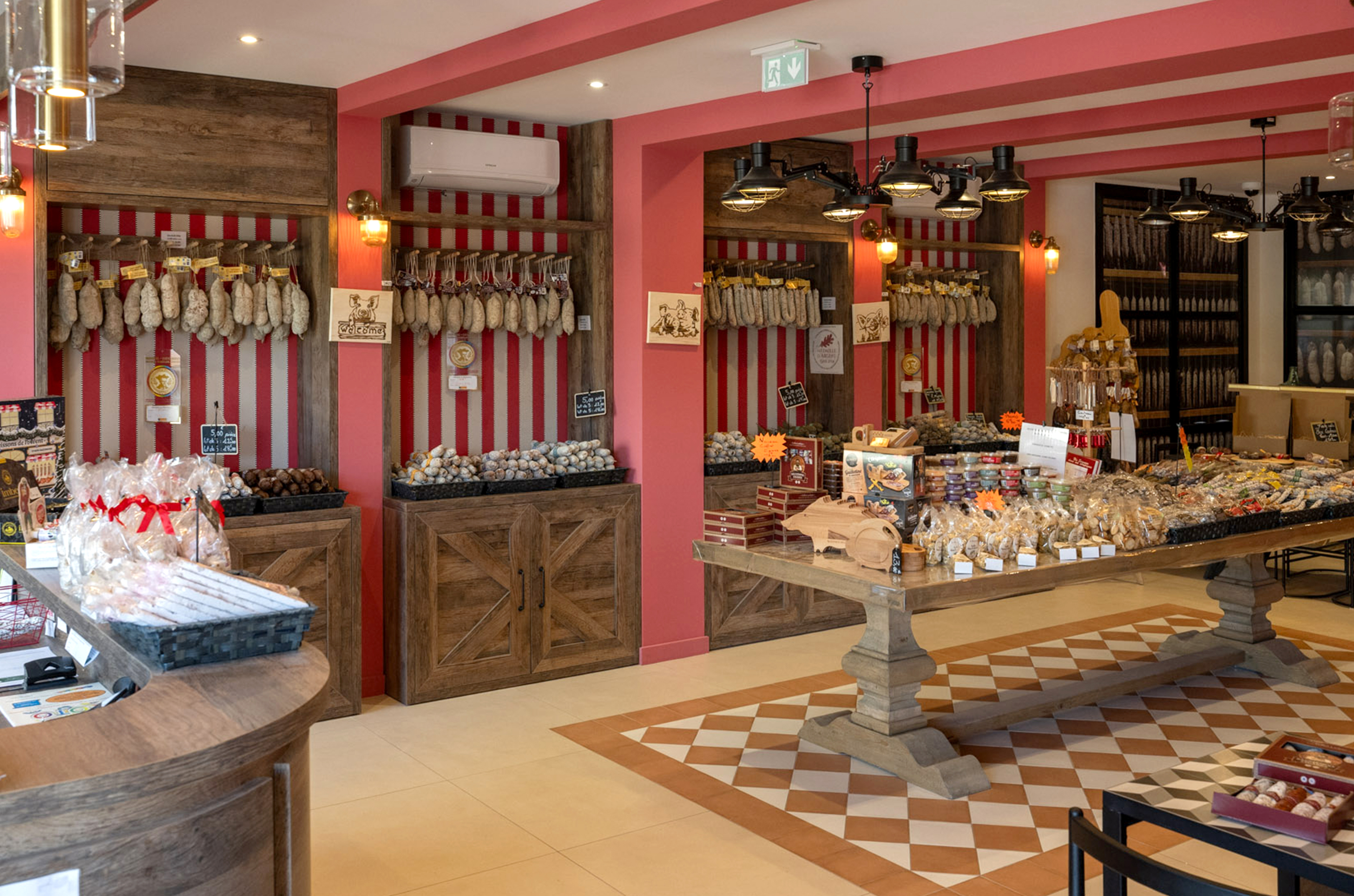Aménagement intérieur d'une boutique de charcuterie par Studio Vesta, avec un sol en damier terracotta, des meubles en bois brut et des murs à rayures rouges évoquant les boucheries traditionnelles.