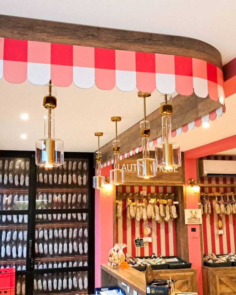 Mise en valeur des produits par un éclairage soigné avec des suspensions cylindriques dorées sous une marquise à festons rouges et blancs.
