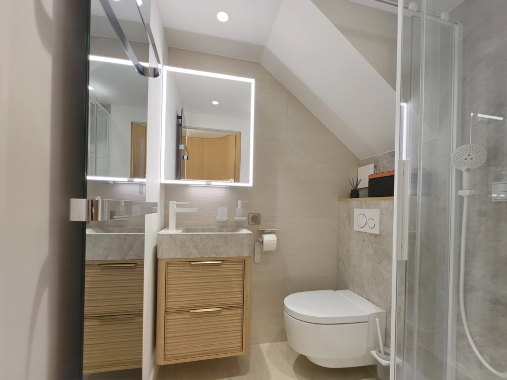 Salle d'eau compacte sous pente avec douche, WC suspendu japonais, meuble vasque en bois strié et miroir rétroéclairé.