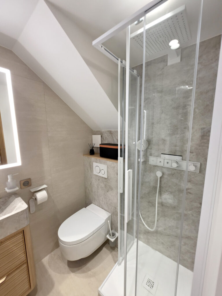 Douche à l'italienne sous pente avec paroi vitrée, robinetterie encastrée blanche et WC suspendu lavant.