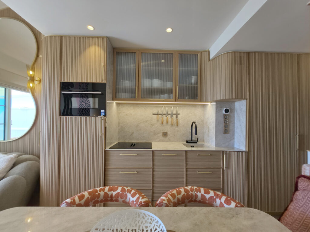 Cuisine compacte sur mesure intégrée sous mezzanine avec façades en bois strié, vitrines armées, crédence marbre et robinetterie noire.