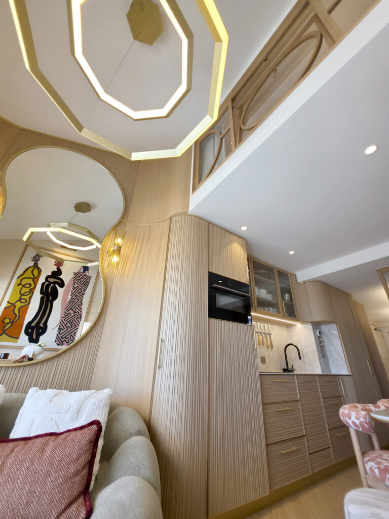Vue en contre-plongée du salon montrant la cuisine en bois strié, le lustre géométrique doré et la verrière de la mezzanine.