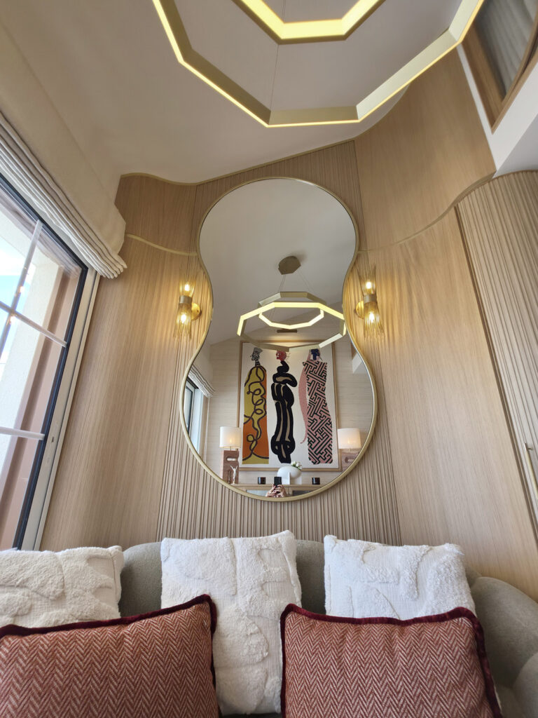 Grand miroir organique au cadre doré reflétant l'œuvre d'art des trois femmes et la suspension lumineuse hexagonale.