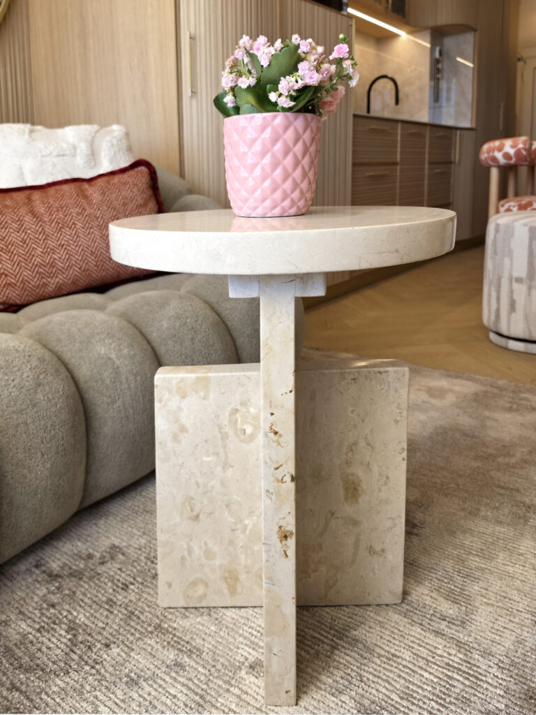 Table basse d'appoint circulaire en marbre/travertin clair avec vase rose géométrique à côté du canapé capitonné.