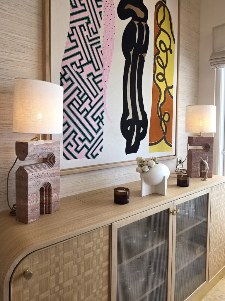 Plateau du buffet en bois clair avec lampes à poser en travertin rouge sculpté et bougies parfumes sous l'œuvre textile colorée.