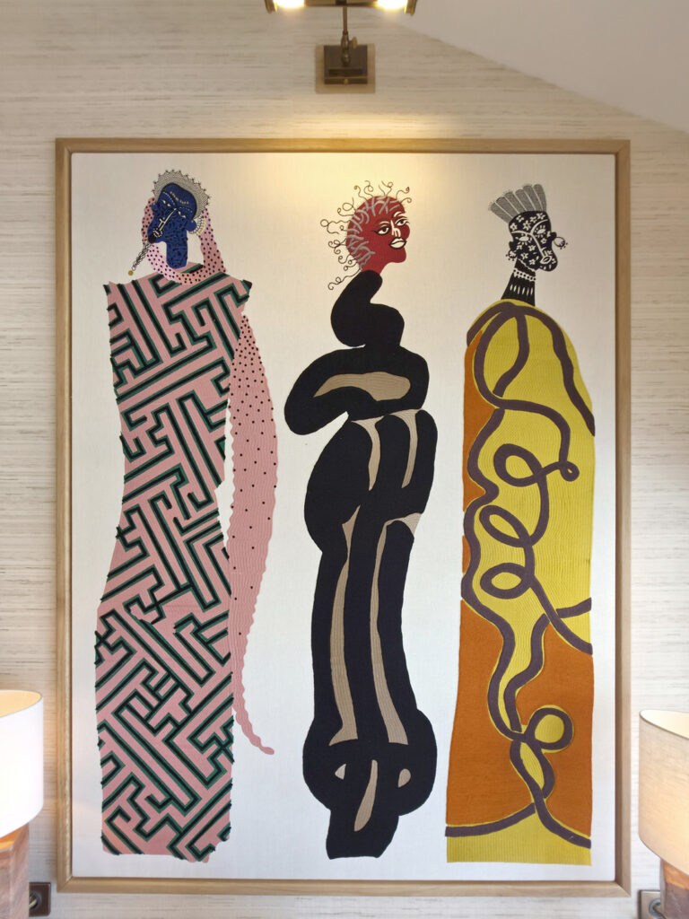 Tableau textile d'art monumental représentant trois silhouettes féminines stylisées aux motifs géométriques et teintes terracotta, ocre et rose.