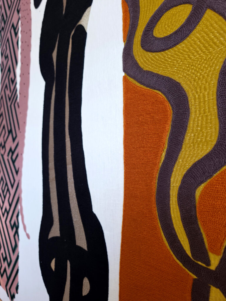 Gros plan sur les textures de broderie et de tissage du tableau d'art, révélant les reliefs des fils terracotta, jaunes et noirs.