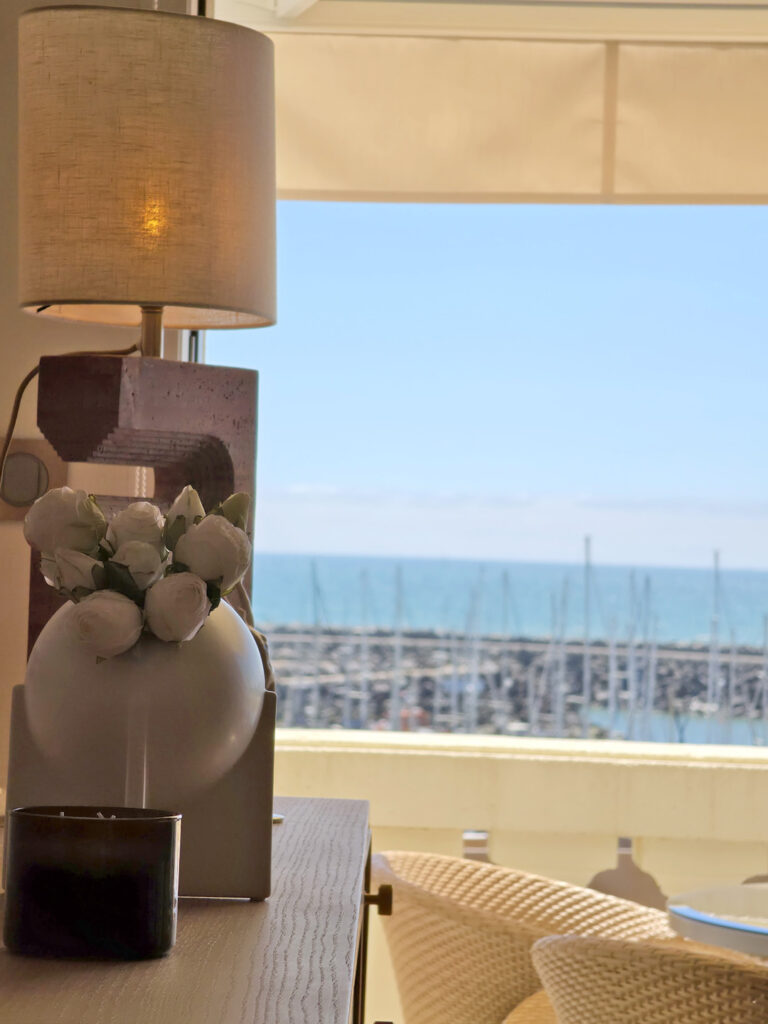 Lampe à poser en travertin et vase en céramique blanche sur le buffet, avec le port de Bourgenay en arrière-plan flou.