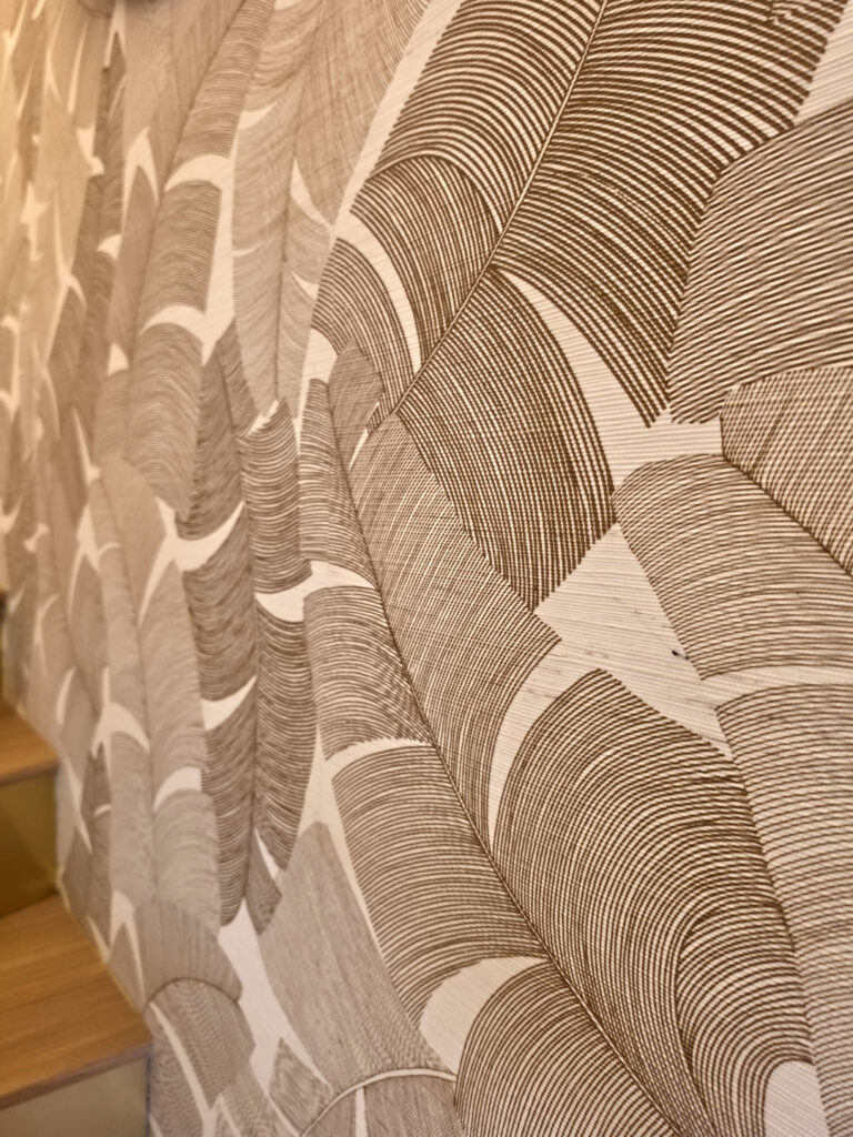 Gros plan sur la texture gravée et tissée d'un papier peint panoramique motif palmes tropicales aux teintes beige et bronze.