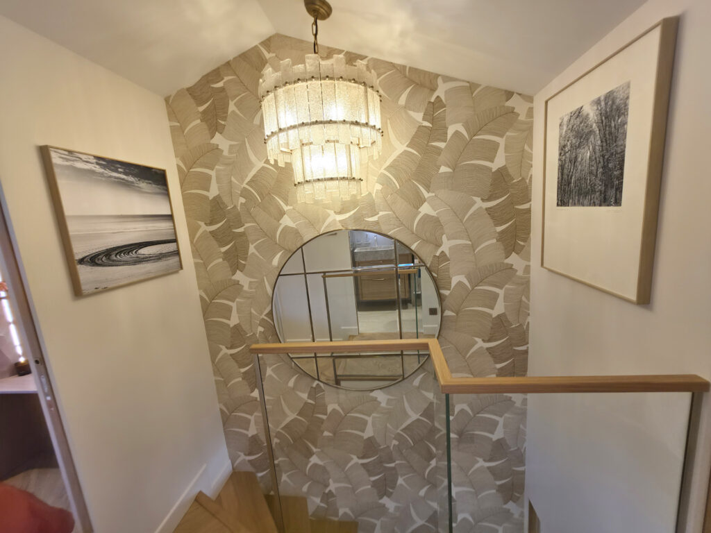 Cage d'escalier vue du palier avec garde-corps en verre, miroir circulaire à parecloses, papier peint palmier et lustre cascade en laiton.