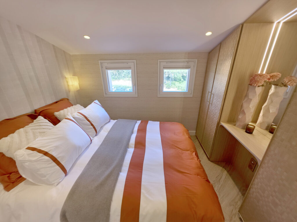 Vue grand angle de la chambre parentale mansardée avec deux fenêtres, grand lit coffre terracotta et dressing sur mesure éclairé.