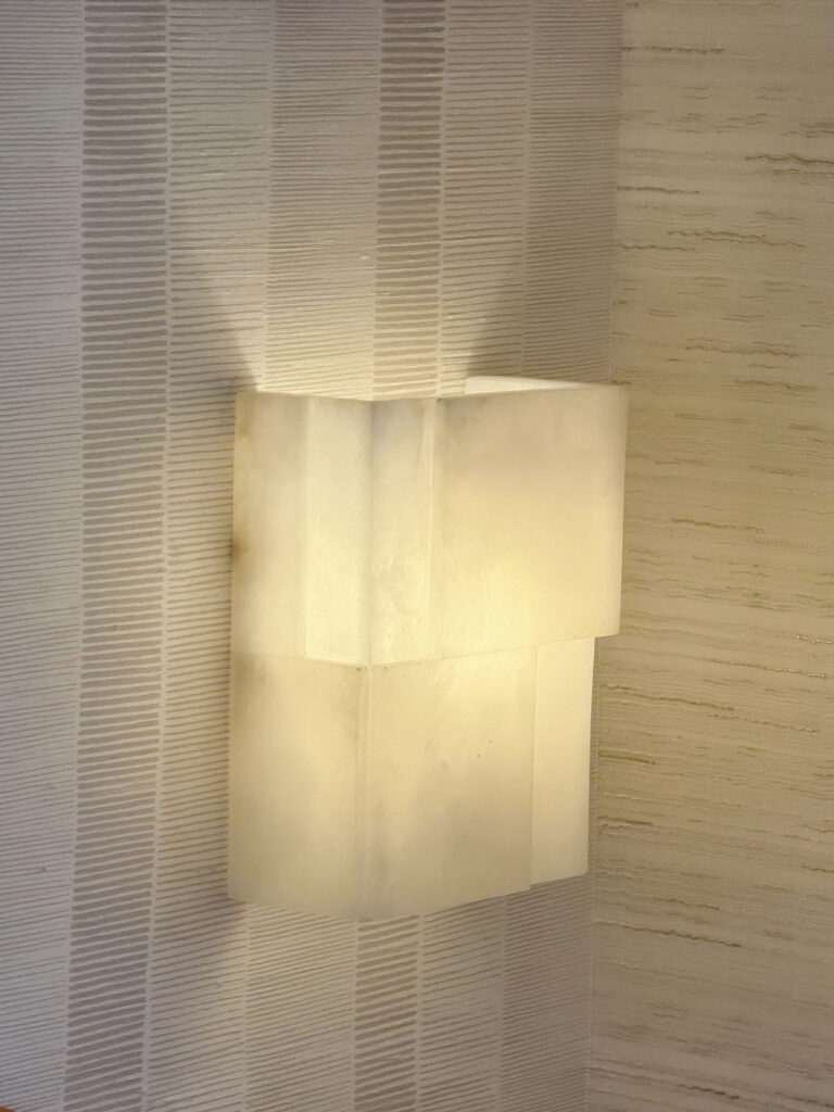 Applique murale architecturale en albâtre diffusant une lumière chaude sur papier peint texturé beige.