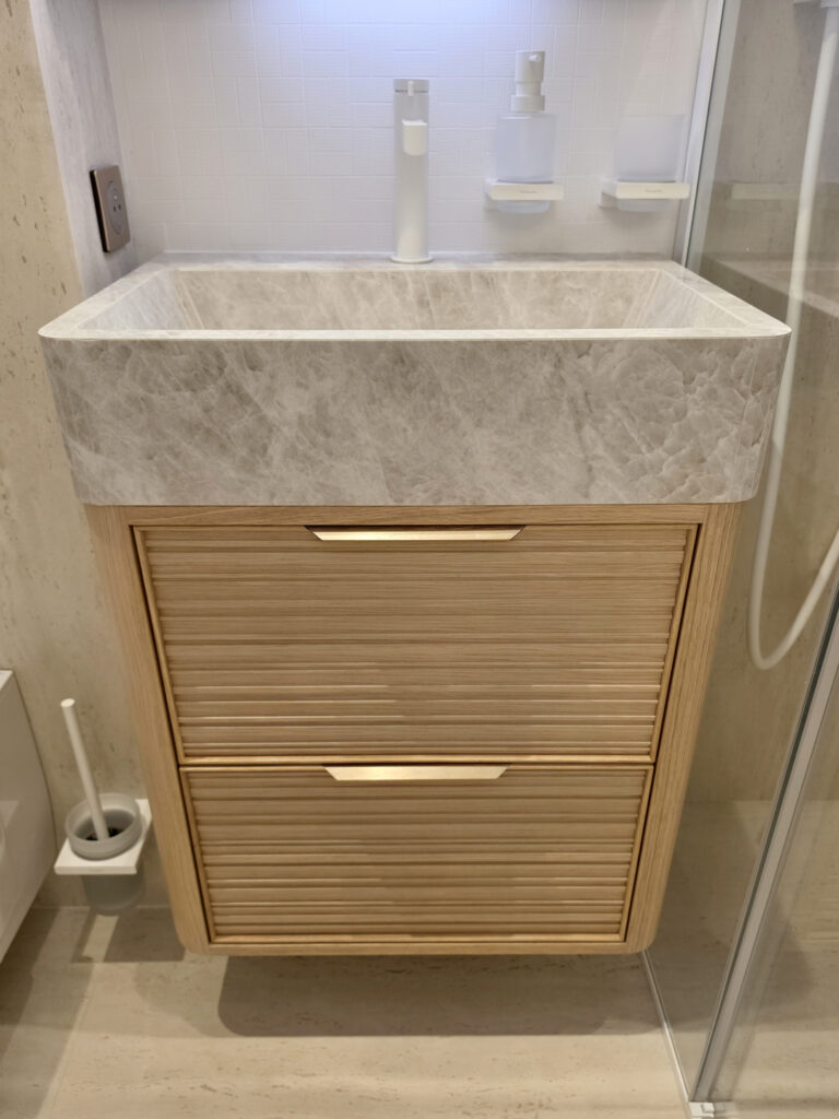 Meuble vasque sur mesure en chêne clair strié surmonté d'une vasque taillée dans du marbre veiné beige avec robinetterie blanche.