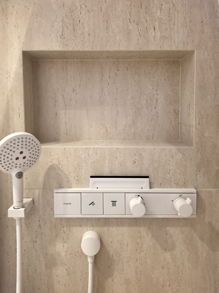 Robinetterie de douche Hansgrohe encastrée blanche thermostatique sous une niche à produits intégrée dans le carrelage effet travertin.