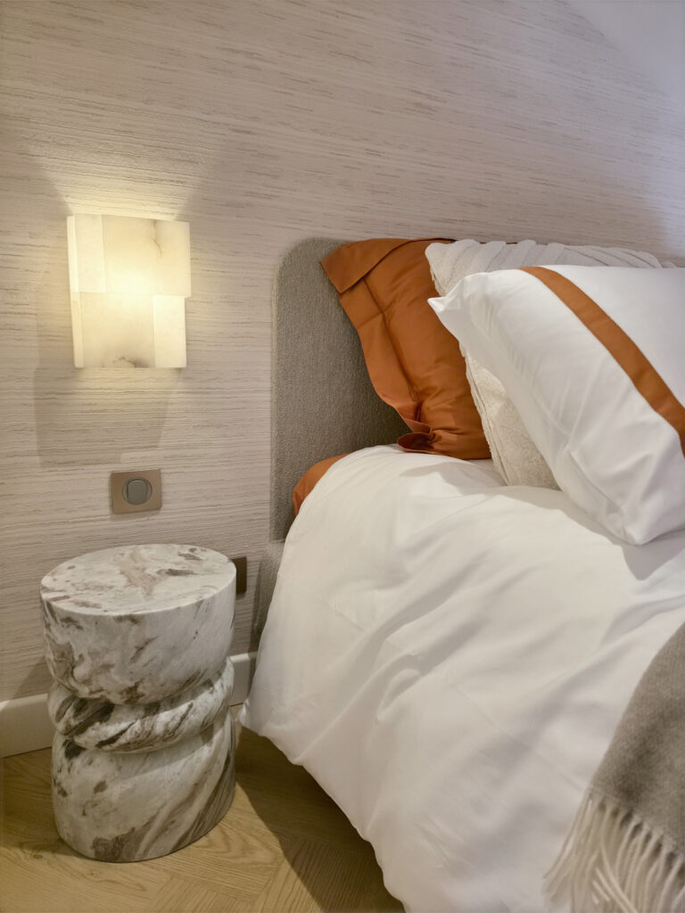 Tête de lit grise, lit coffre avec couette terracotta, applique en albâtre et table de nuit sculptée en marbre veiné.