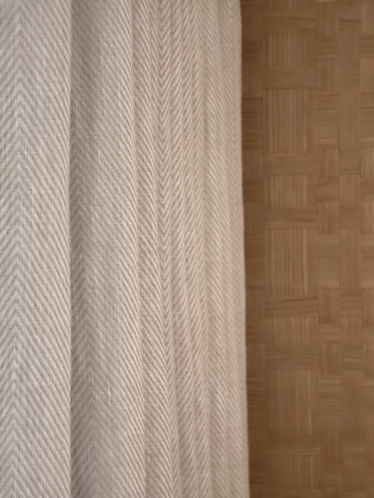Gros plan sur la rencontre des matières entre le tissu chevronné lin des rideaux et le panneau d'ébénisterie en bois tressé.