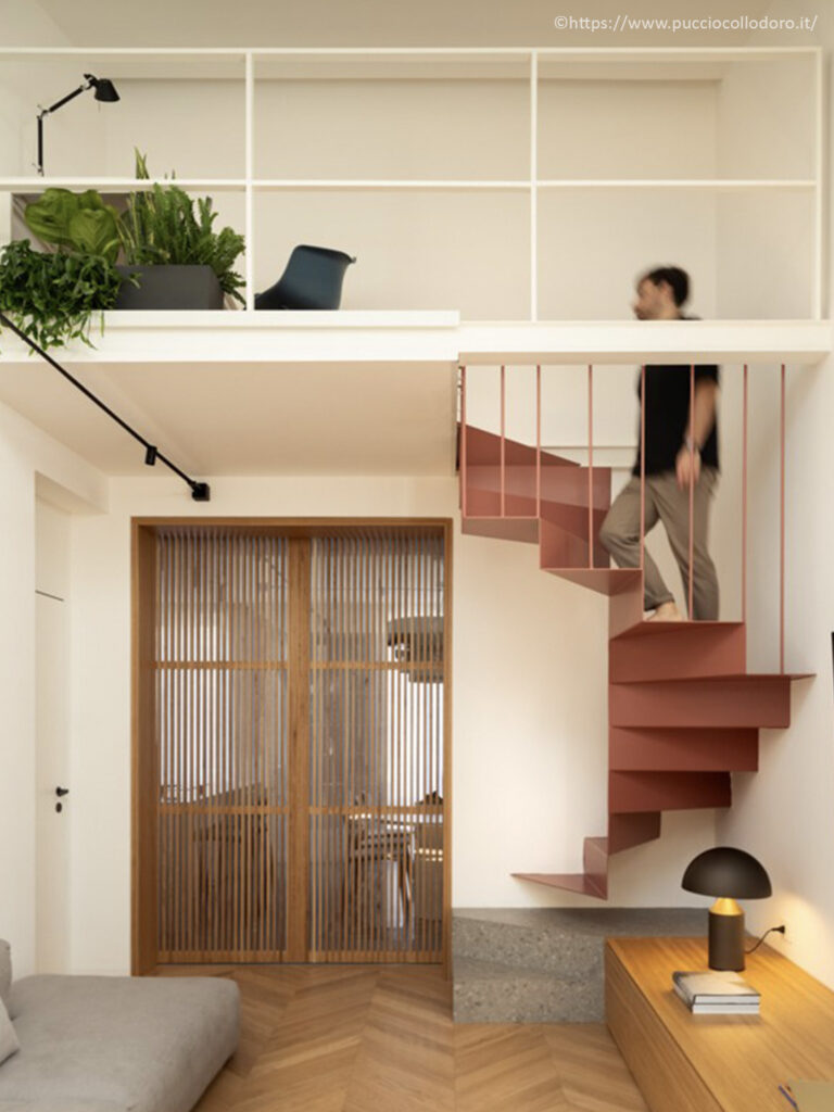 Escalier hélicoïdal suspendu couleur terracotta menant à une mezzanine nuit sous plafond blanc par PuccioCollodoro Architetti.